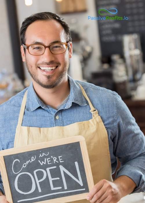 A photograph of a small business owner wearing an apron, smiling, and holding a sign that says come in we're open.